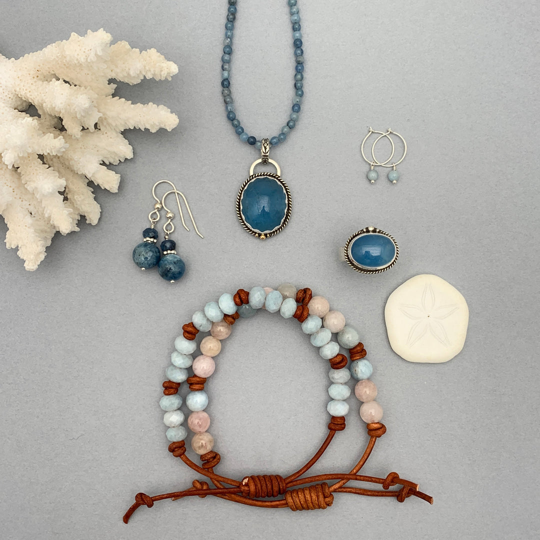 Blue beaded necklace with aquamarine gemstone pendant, aquamarine bead and leather bracelets, silver hoop earrings with aquamarine charm, silver dangle earrings, and aquamarine gemstone ring, on gray background with white coral and sand dollar accents.